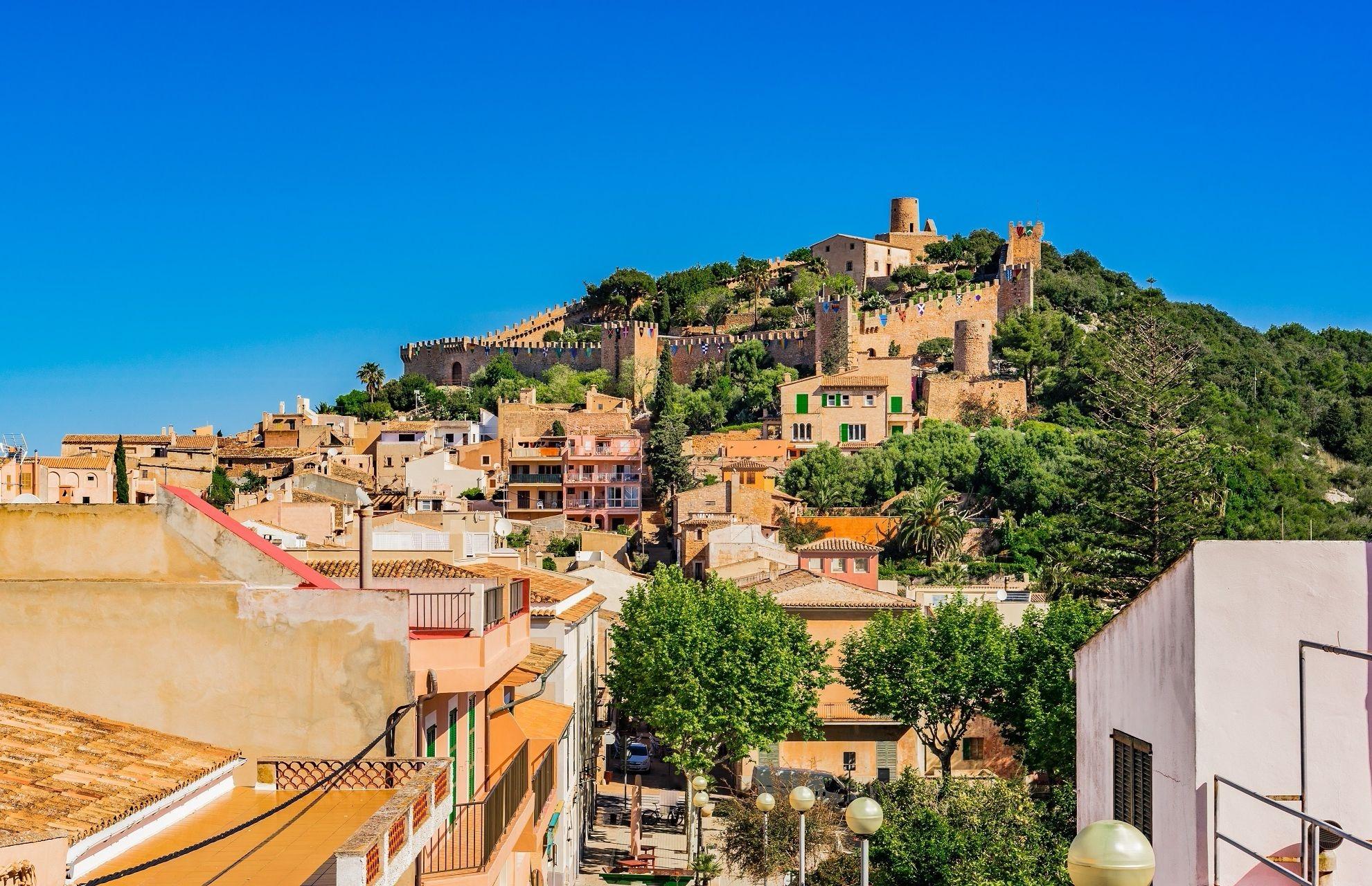 Colorful houses squeeze along a green hilly landscape. At the top, there is a medieval castle with battlements. The peaceful panorama of this Mediterranean scene is draped under a bright blue sky.