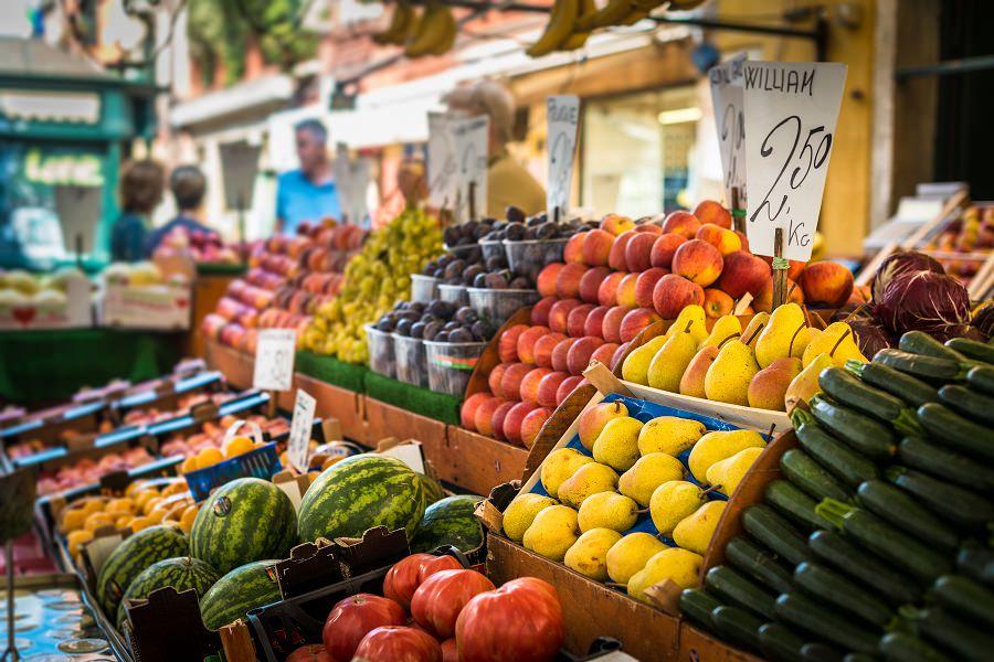 On a market stand, watermelons and tomatoes lie on the left, while mounds of pears, apples, and plums are on the right. All fruits are stacked in wooden boxes, and price tags stick up from the displays.