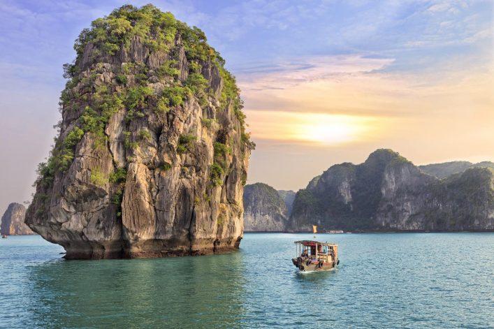 Halong Bay in Vietnam: Majestic limestone cliffs with green vegetation on the left, calm blue water, traditional wooden boat on the right; sunset creates warm light, background of hills framed.