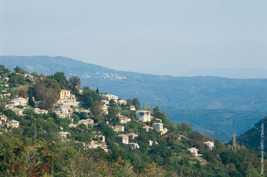 Hills with green vegetation and white houses in Greece. In the foreground trees, in the background a vast hilly landscape under a clear sky.
