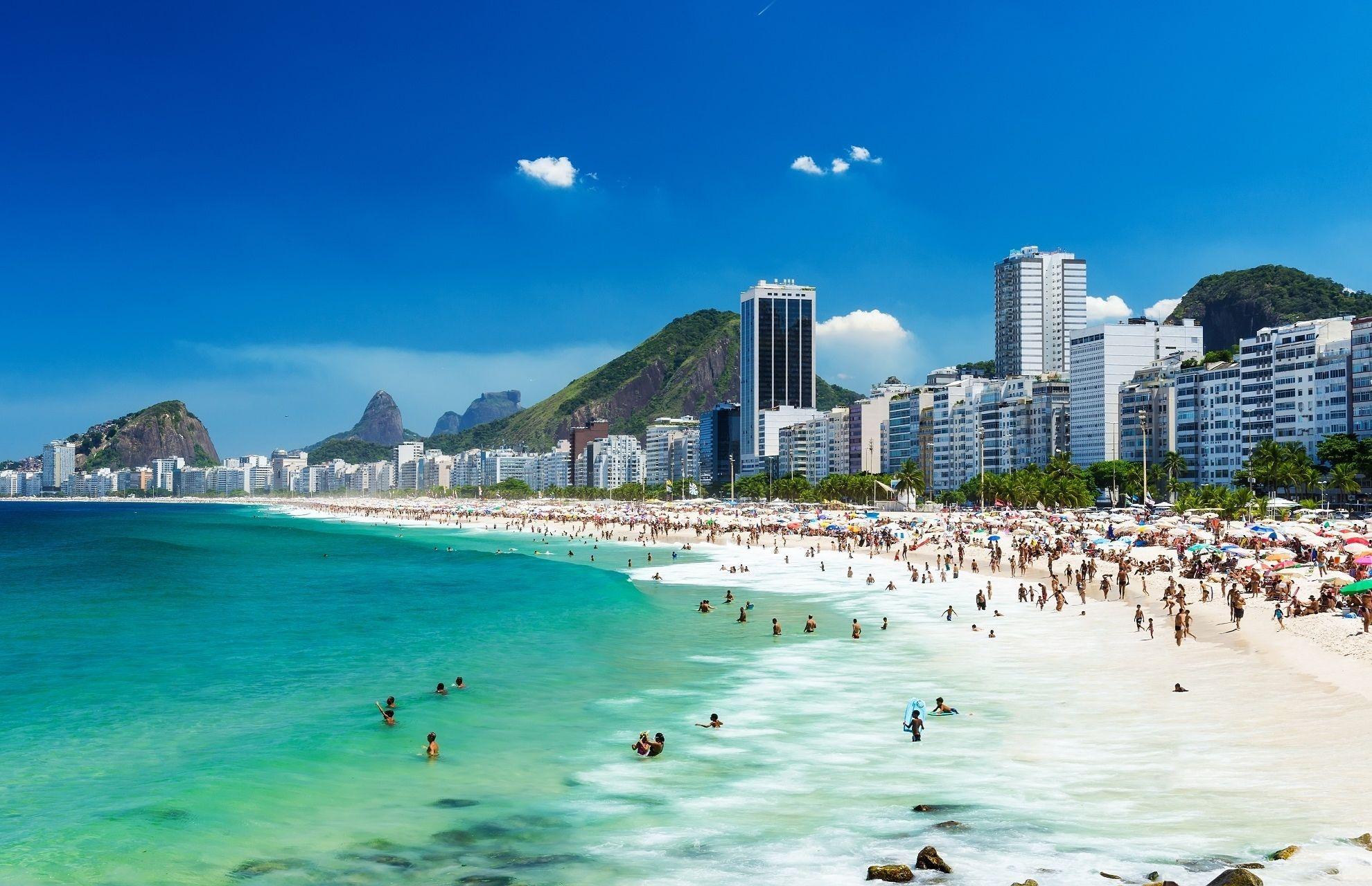 Copacabana in Rio de Janeiro on a sunny day. On the left, the turquoise sea with swimmers. On the right, the wide, white sandy beach filled with people and colorful umbrellas. In the background high-rises and green hills. Copacabana is a popular stop on cruise routes through South America.