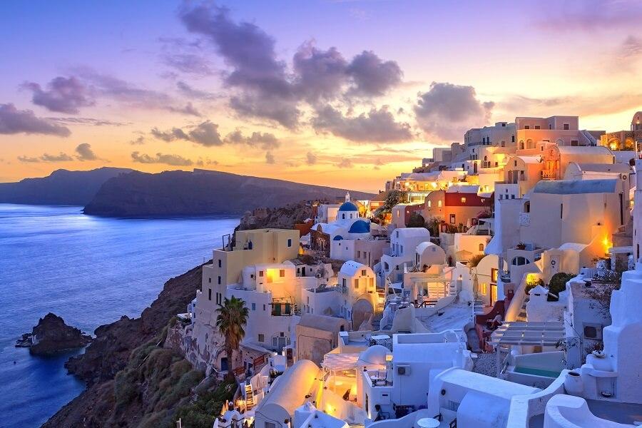 Whitewashed houses with blue domes stretch over a cliff in Santorini, illuminated by the warm light of a sunset. In the background, a calm sea and a distant hill.