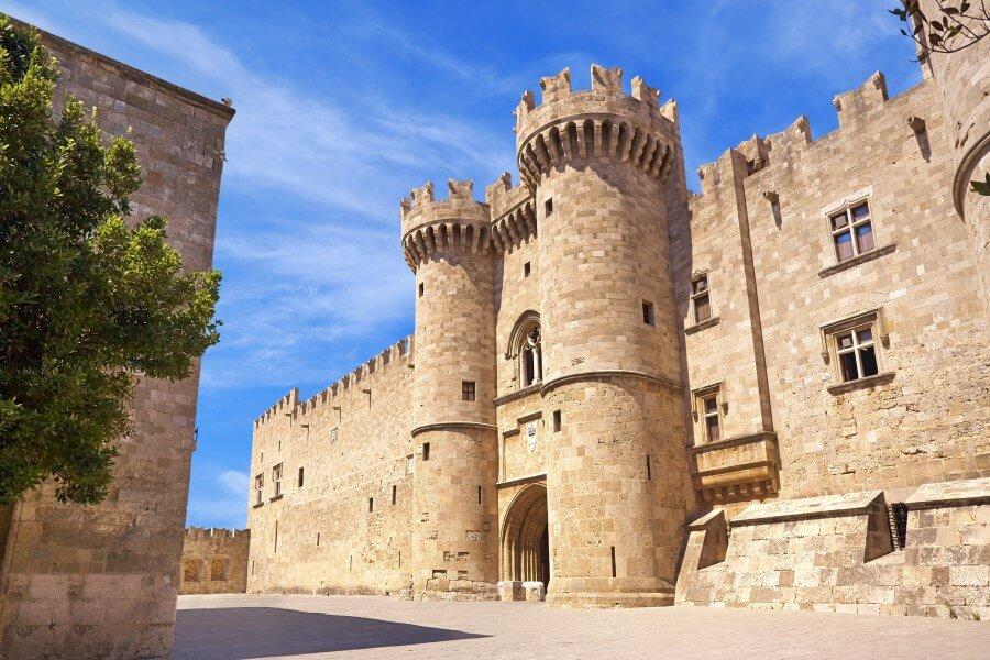 Mighty stone walls of the Grand Master Palace with battlements and two round towers under a blue sky. The central archway looks inviting. On the left, a tree, and on the right, shadows on the ground.