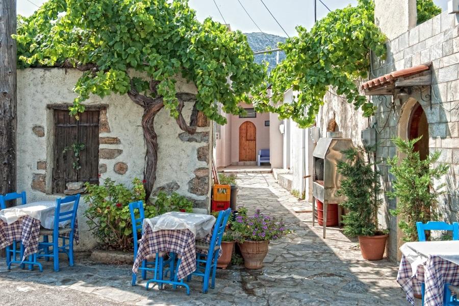 A restaurant in Crete located in a narrow alley. Small tables are set closely together, covered with red and white checkered tablecloths, and two blue-painted wooden chairs face each other at each table. Trees grow high along the house walls, and flower pots with colorful flowers are placed in the alleys.