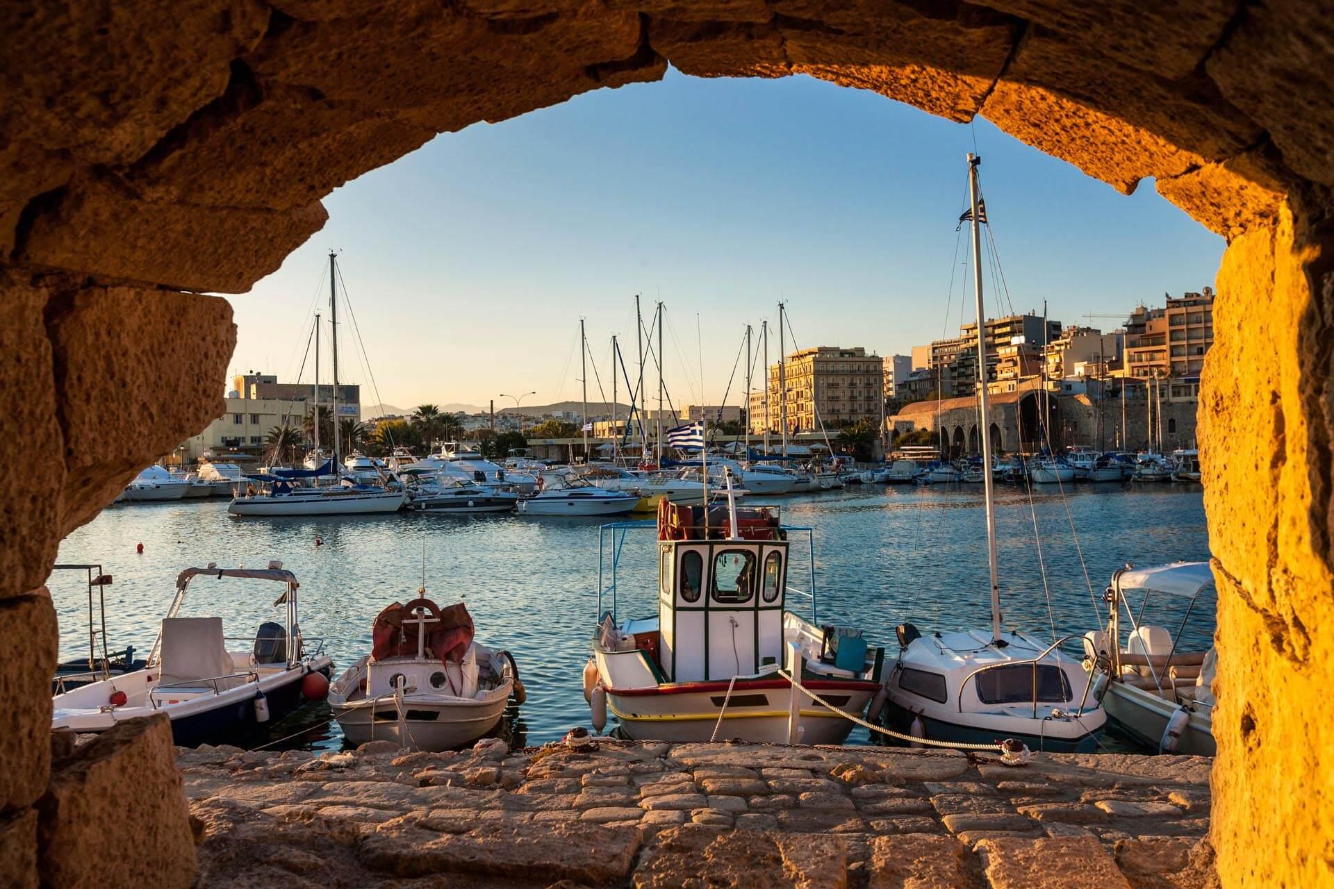A lovely marina in Crete. Photographed through an arch, which can still be faintly seen at the top of the image. The ground is paved with cobblestones, and behind them, small white boats float in the water. The image was taken at twilight, casting a warm yellow-orange light.
