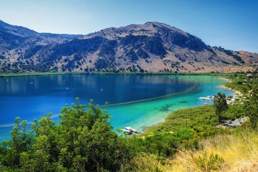 View of Lake Kournas in Crete. It is surrounded by high cliffs. The water is deep blue in the center of the lake and becomes clearer and lighter towards the shore. Colorful pedal boats are lined along the shore. The sky is blue.