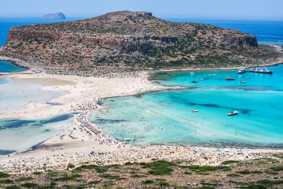 Aerial view of Balos Lagoon in Crete. The lower and upper edges of the image show the mainland or an island. A narrow white sandy beach runs through the center of the image, swarmed with bath tourists. Crystal-clear water can be seen on the left and right of the beach.
