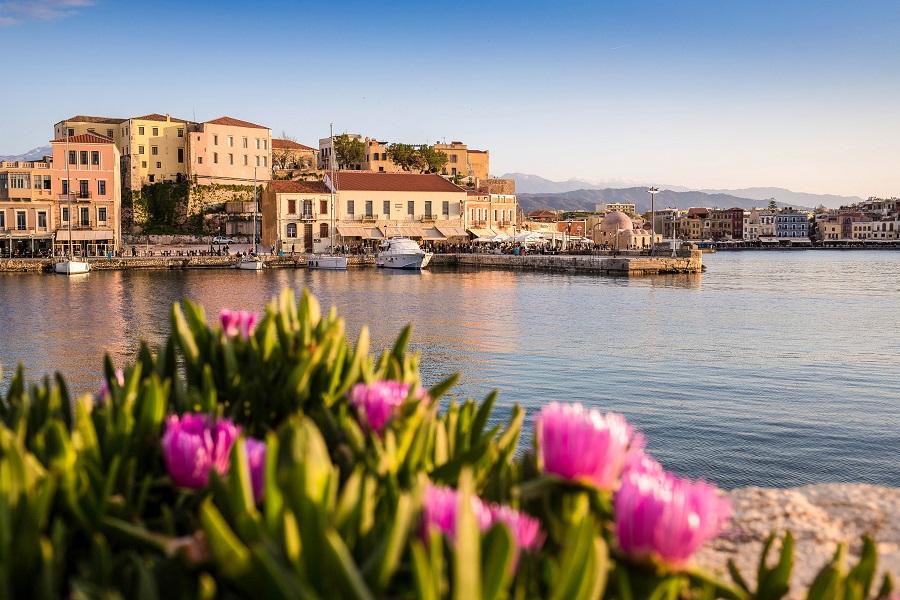 Colorful harbor in Chania with historic buildings in the background. In the foreground, blooming pink flowers, behind calm water and a picturesque sky. Mountains are visible in the distance.