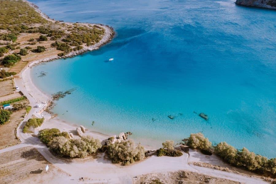 Aerial view of Voulisma Beach in Crete. The majority of the picture showcases the turquoise water. On the left edge, the coast can be seen, partially covered with green bushes. The beach, empty in the image, reveals white sand lying between the coast and the water.