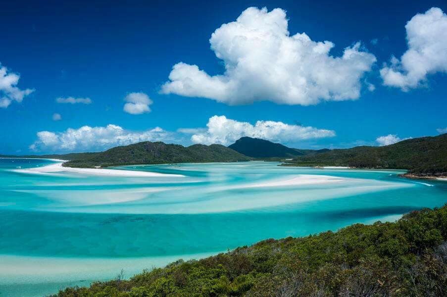 Turquoise sea with white sandbanks stretches before green hills. White clouds float in the sky. Clear, sunny atmosphere.