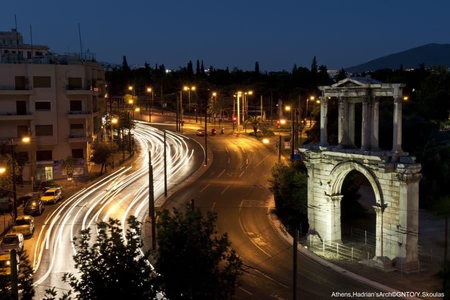 At night, car trails glow on a winding road. On the right stands the illuminated Hadrian's Gate. On the left is a building, with trees and the deep blue sky in the background.