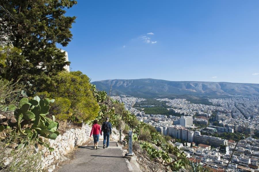 Two people walking on a path on Lycabettus Hill, with a view of Athens in the background. Trees and cacti grow on the left, while the city stretches out under a clear sky on the right.