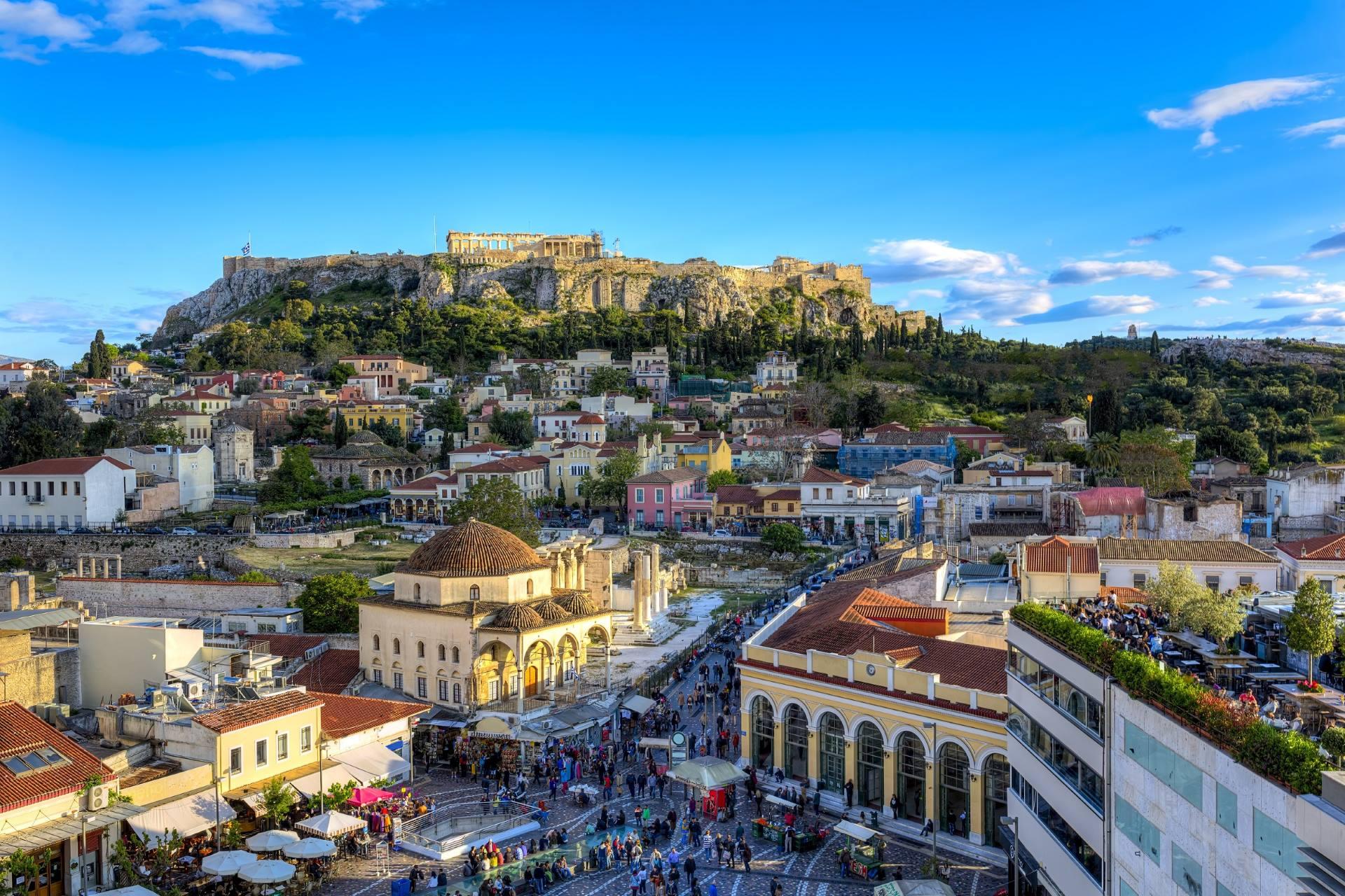 Image of the Acropolis in Athens. The lower part of the image shows a city with houses, people, and scattered trees. In the background lies the Acropolis. The sky is blue.