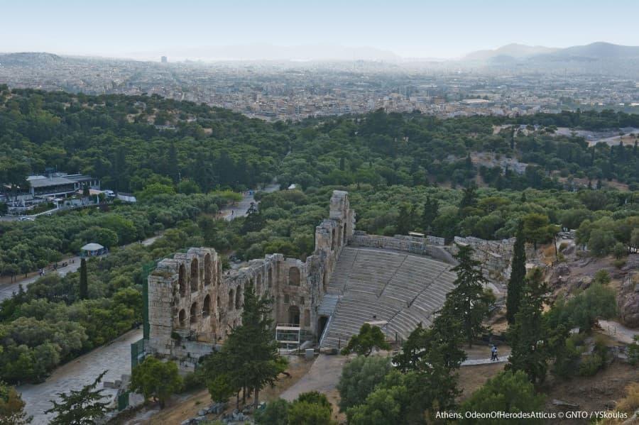 View of the Odeon of Herodes Atticus in Athens, an ancient stone theater surrounded by trees and hills. In the background, the city of Athens stretches to the horizon.
