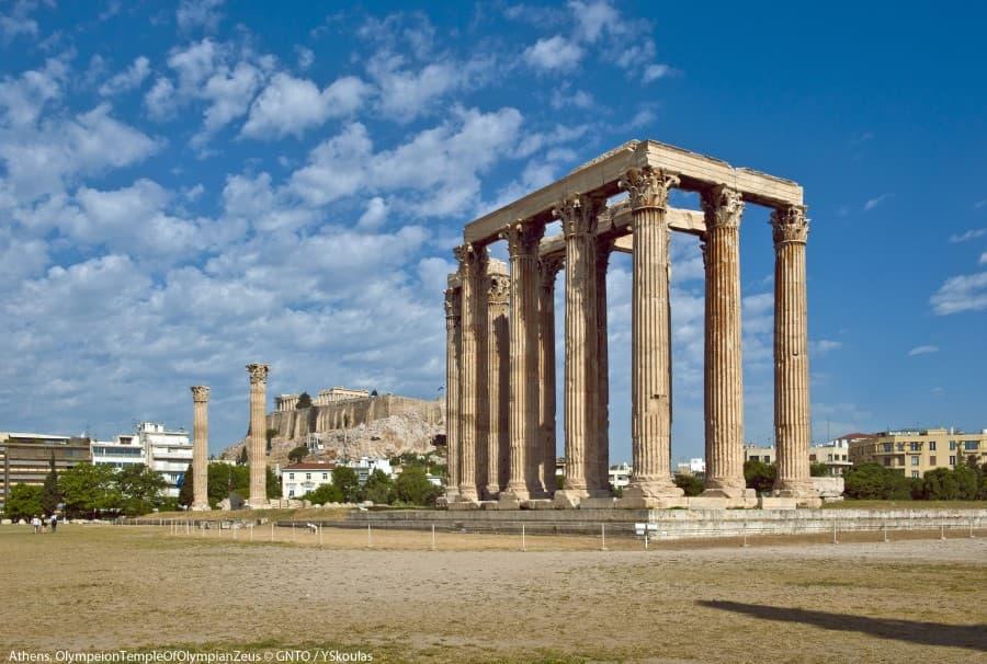 Ancient columns of the Olympeion Temple in Athens rise majestically into the blue sky. In the background, the Acropolis extends, surrounded by modern buildings and green grass in the foreground.