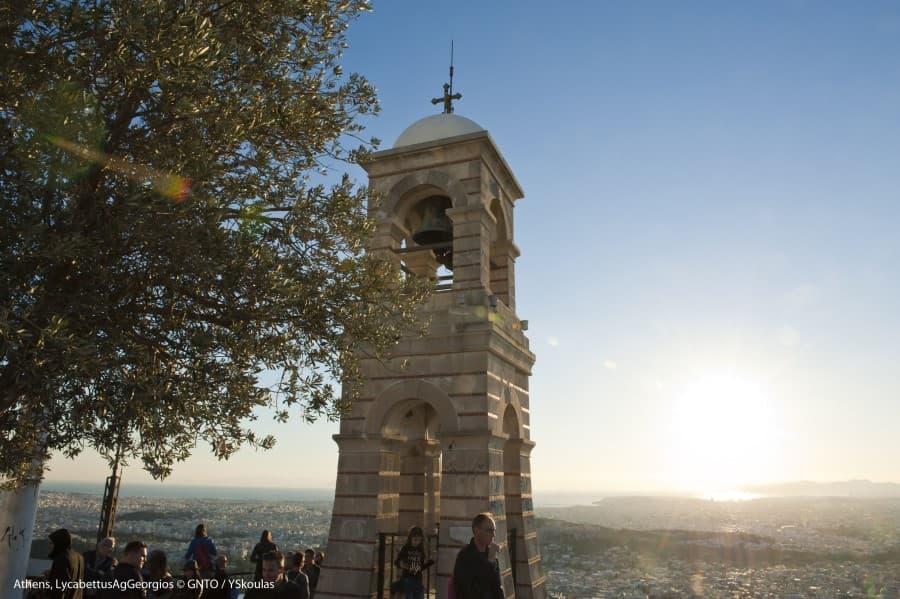 Tower with cross under the blue sky; sunset in the background. People stand around the tower, partly covered by a tree. View of a vast city landscape.