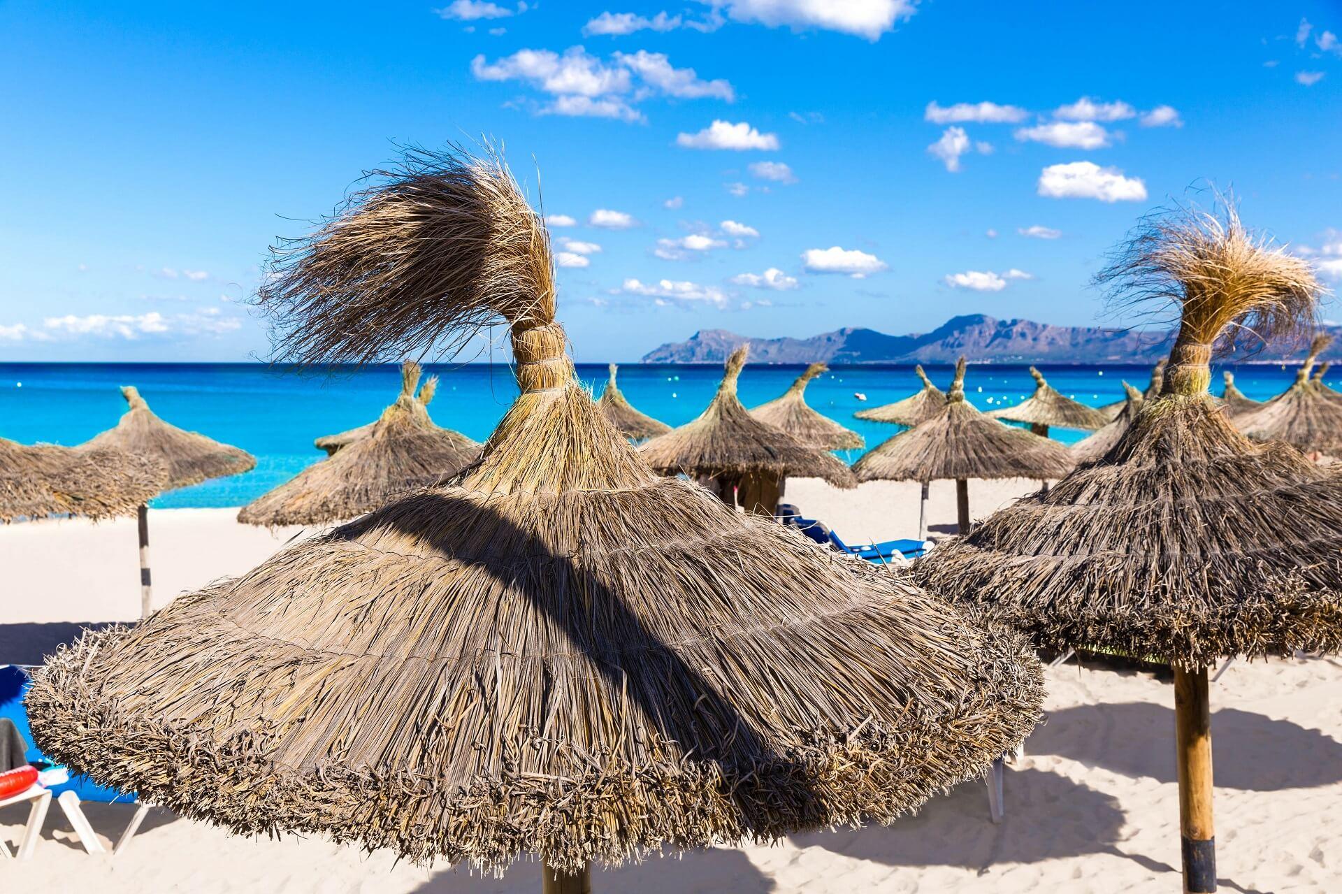 The beach of Can Picafort, focusing on round straw beach umbrellas. The sky is blue and the clear sea shimmers on the horizon.