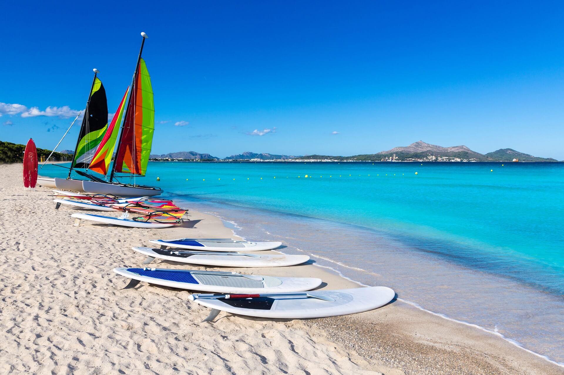 Platja del Muro in Mallorca. The water shimmers in bright and clear blue, the sky is the same color, and white surfboards and boats with colorful sails are laid out on the white sandy beach in the foreground.