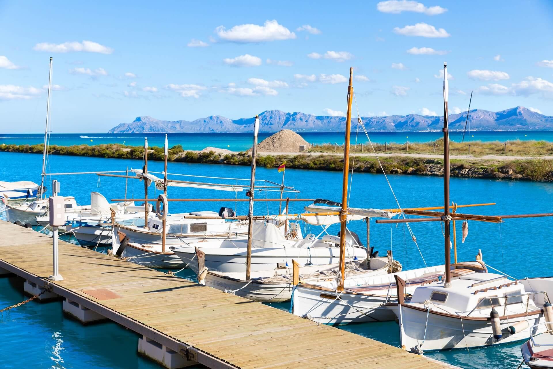 A wooden walkway leads into the blue water at Platja del Muro. In front of the walkway, some small white boats lay anchored. Behind them, a narrow, green-covered peninsula juts into the sea, with mountains visible in the background. The sky is blue with a few clouds.