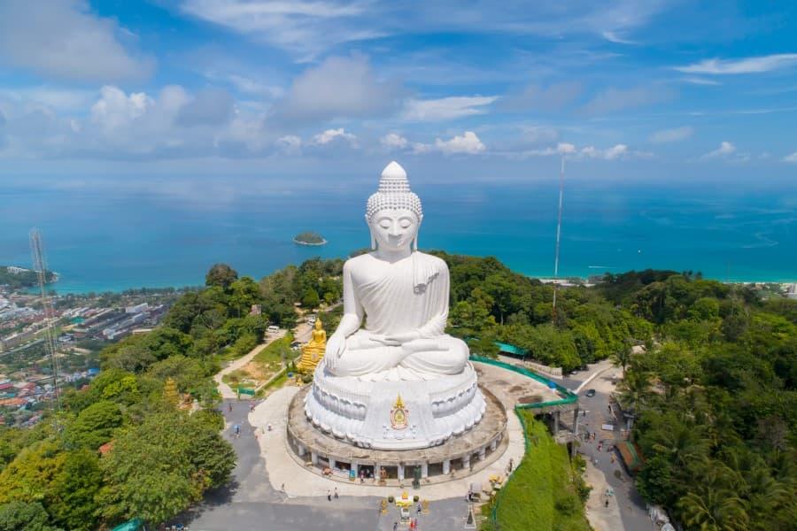 The gigantic white Big Buddha of Phuket majestically rises on a hill with lush greenery in front. In the background stretches the turquoise sea as a popular cruise route towards Thailand.