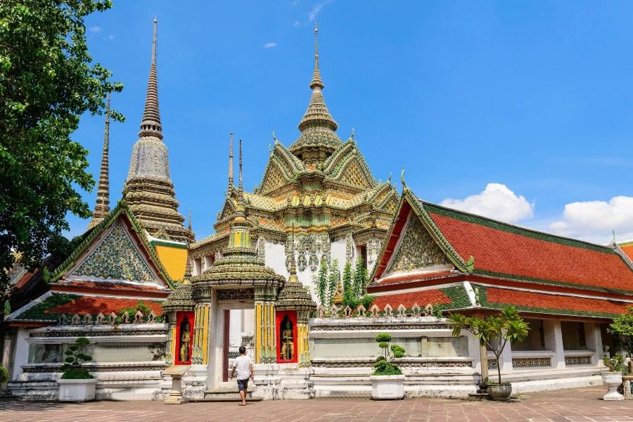 Magnificent temple in Bangkok with intricate roofs and tall, ornate towers. A visitor walks through an archway. On the left is a tree, and on the right is a small palm shrub. Blue sky.