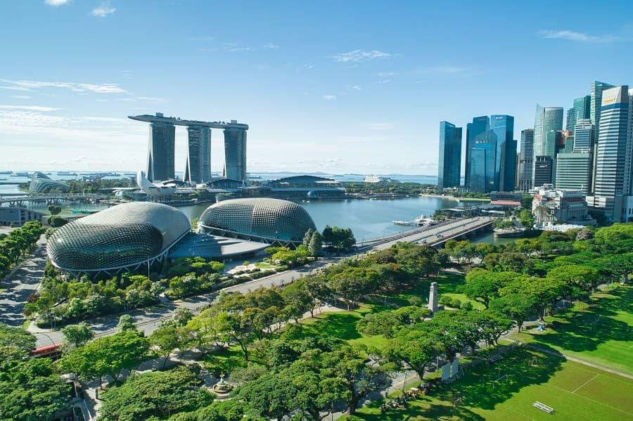 A green park with trees in the foreground, with modern buildings and bridges behind, on the right the skyline of Singapore. In the background, the distinctive Marina Bay Sands hotel with three towers.