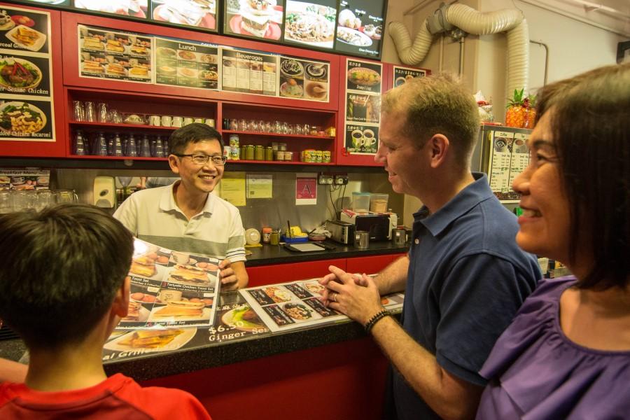 A vendor in a hawker center smiles as he talks to three people at a counter. In the background, menu boards and photos are visible on the wall.