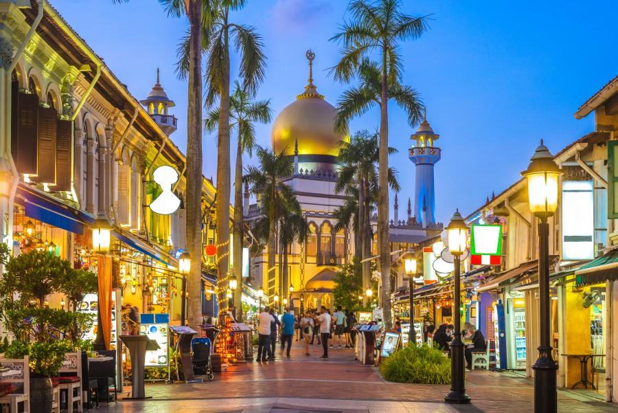 Evening atmosphere in Singapore, Kampong Glam: The Sultan Mosque with a golden dome in the background, flanked by palm trees and busy, illuminated shops on both sides of the sidewalk.