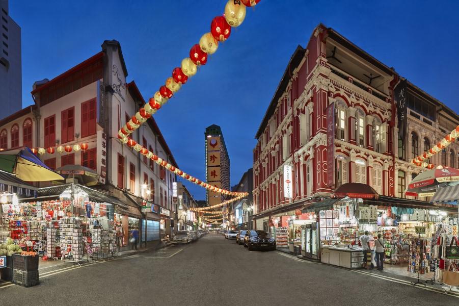 Chinatown in Singapore at dusk: Colorful lanterns hang over a busy street with historical red-and-white buildings and market stalls full of various goods on both sides.