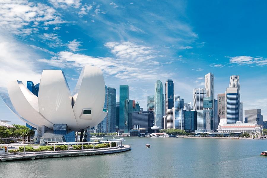 Modern skyline of Singapore with the distinctive petal-shaped ArtScience Museum on the left foreground, surrounded by water. Skyscrapers rise into the blue sky in the background.