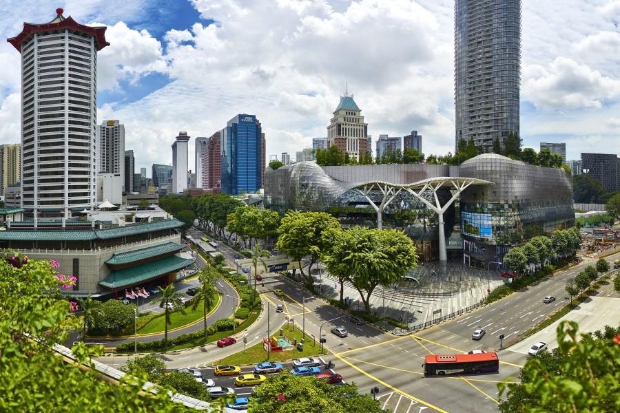 Skyscrapers and modern buildings line Orchard Road in Singapore. Cars and buses move below through complex roads. Trees and green spaces break up the urban scene.