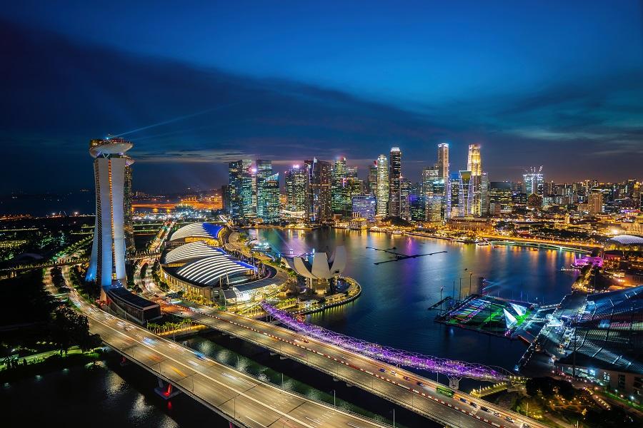 Singapore's night skyline with illuminated skyscrapers, Marina Bay Sands on the left and water in the foreground. A bridge spans the bay, reflecting lights in the water.
