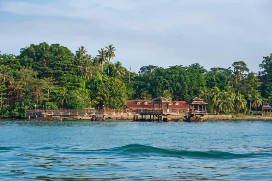 Tropical island with dense green forest in the background. In the foreground, a traditional wooden building on stilts by the water. The sky is slightly cloudy, the sea calm.
