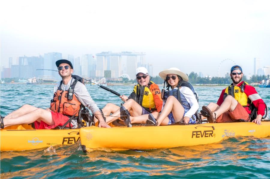 Four people sit in yellow kayaks on the water, wearing life jackets. In the background, the skyline of Singapore and the giant Ferris wheel are visible. The atmosphere looks relaxed and cheerful.