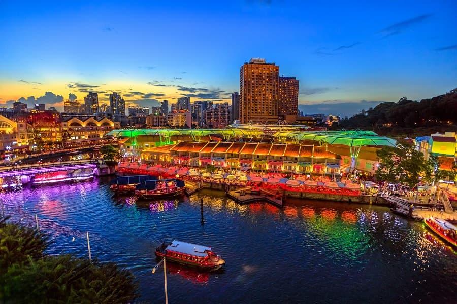 Colorful lights on buildings reflecting in the water of a river at dusk. In the foreground, boats are moving, and in the background, illuminated skyscrapers and a dark sky are visible.