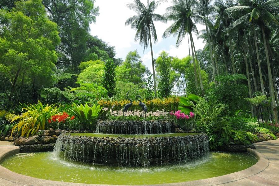 Lush garden with palms in the background. In the center, a multi-tiered fountain surrounded by colorful flowers and dense foliage. Water cascades over the edges of the fountain.