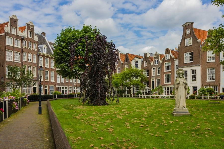 The Begijnhof Amsterdam: Surrounding a green courtyard where grass and some trees grow, slender stone houses are lined up. In the courtyard stands a stone statue.