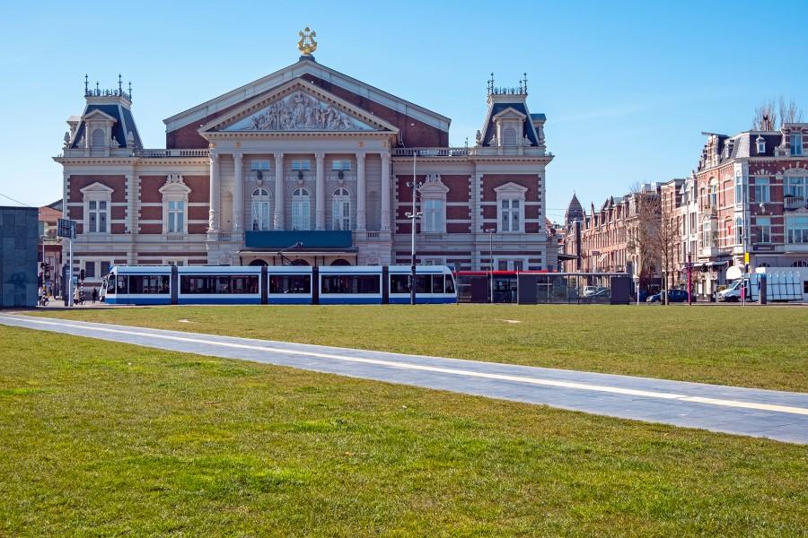 the Koninklijke Concertgebouw in Amsterdam is a classical brick building with classic white decorative elements like protruding columns. A tram passes in front of the building, and a well-kept lawn is in the foreground.
