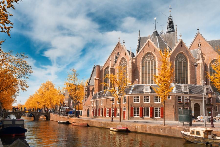 The Oude Kerk in Amsterdam during autumn. The church has an unusual shape and is built in a mix of styles. Bricks and large windows dominate the appearance, with the baroque church tower in the background. In the foreground is a canal, and you can see orange-leaved trees.