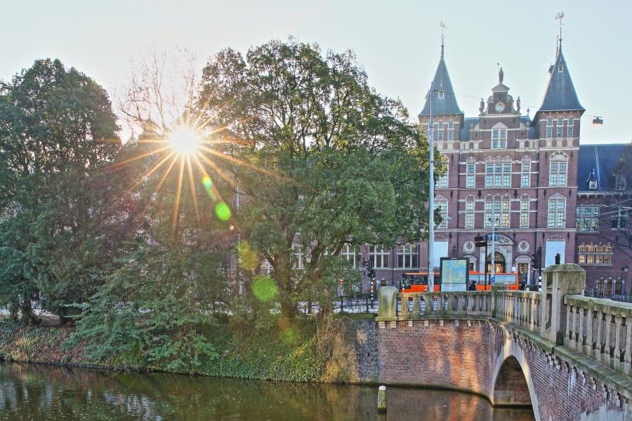 The World Museum in Amsterdam: Sun shines through tree branches, illuminating the walkway and canals of Amsterdam, with a historical building with twin gables in the background.
