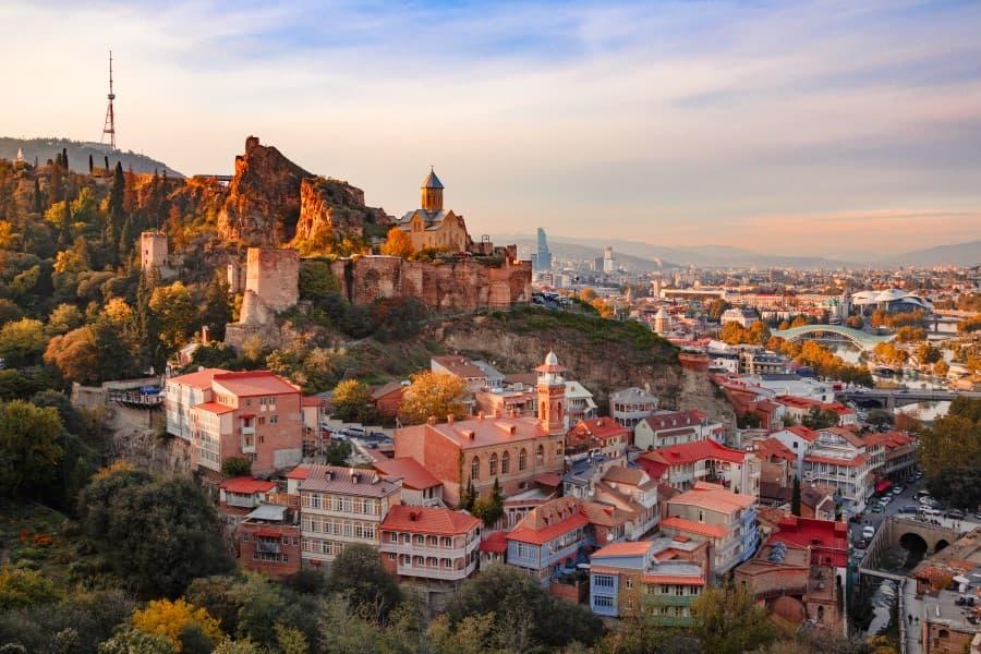 Aerial view of Tbilisi, the capital of Georgia. The fortress looms high atop a mountain overlooking the city. Beneath, many houses with red roofs are visible. In the distance, the Bridge of Peace spans over the river. The photo was likely taken at dusk as the sky is gradually turning purple, and the light is warm yellow tones.