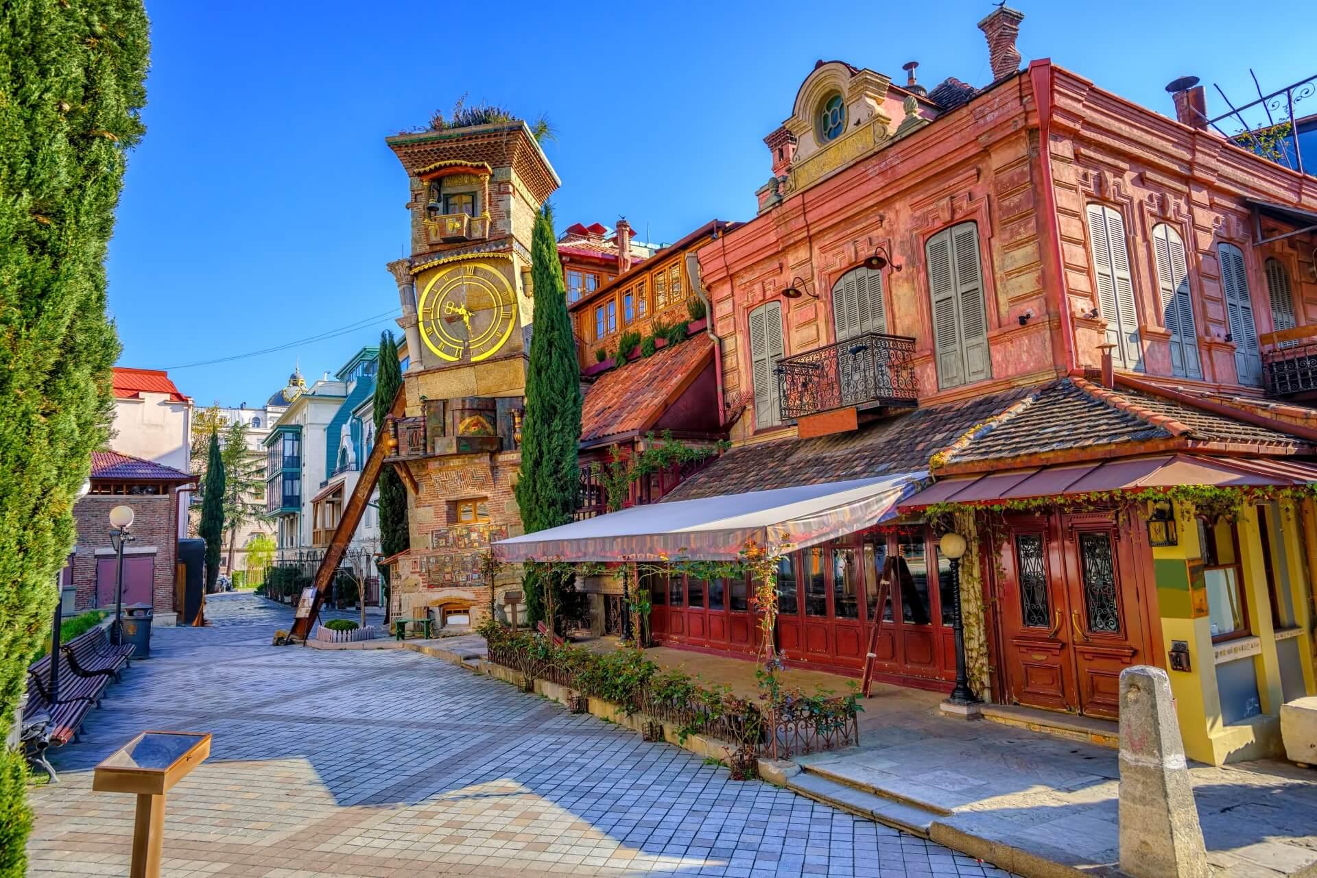 The old town of Tbilisi in Georgia features its old houses. In this image, a red house stands, beside which a slate tower rises high. A large clock is mounted on the tower.