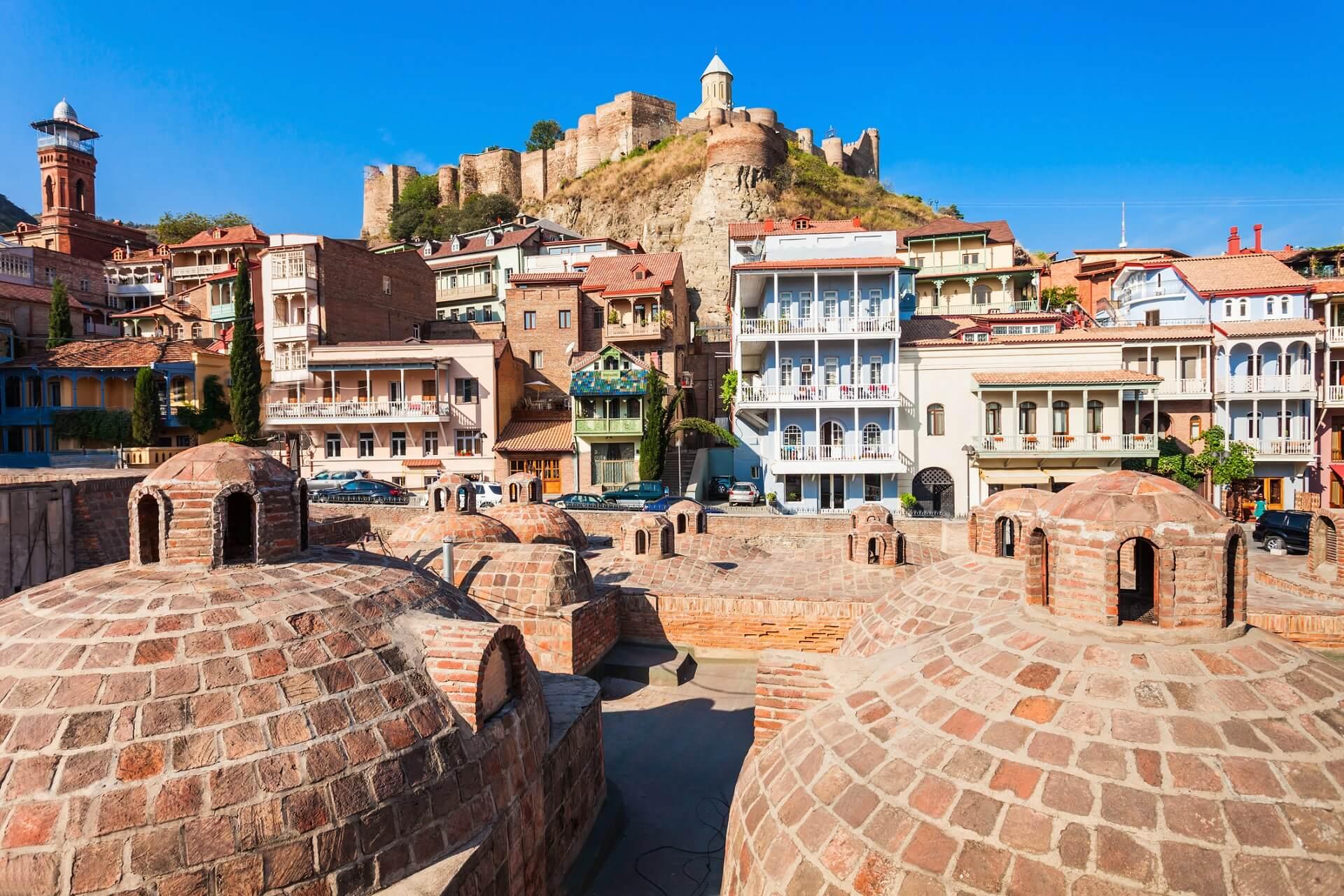 Baths in Tbilisi, the capital of Georgia. The round domes, built from red stones, rise up from the ground in the foreground. In the background, the fortress on the mountain overlooking the city is visible.