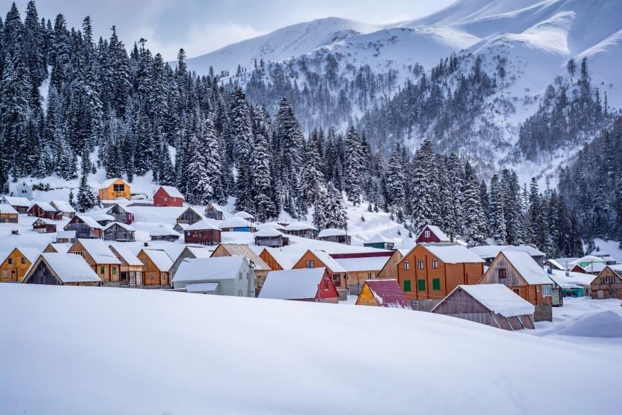 Winter landscape in Georgia. In the foreground, there is a hill covered with snow. Behind it, many colorful houses stand out against the white landscape. Behind the houses are snow-covered fir trees on one side, and high mountains loom in the background.