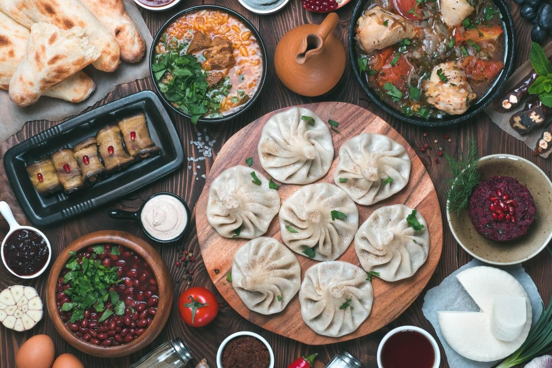 Georgian specialties are freshly served on the plate here. A richly set table is photographed from above. In the middle lies a wooden board on which round-shaped dumplings are placed. Around it are bowls with sauces, dips, and other delicacies.