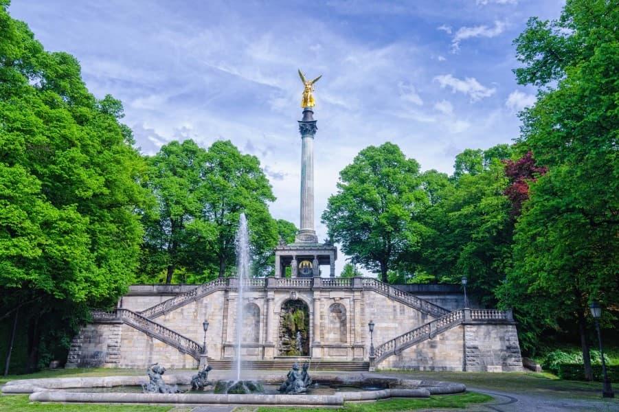 Vista da Estátua do Anjo da Paz em Munique. A estátua dourada está em uma alta coluna, que se destaca sobre as árvores verdes do lado esquerdo e direito. No centro, em frente à coluna, há uma fonte com um jato d'água.