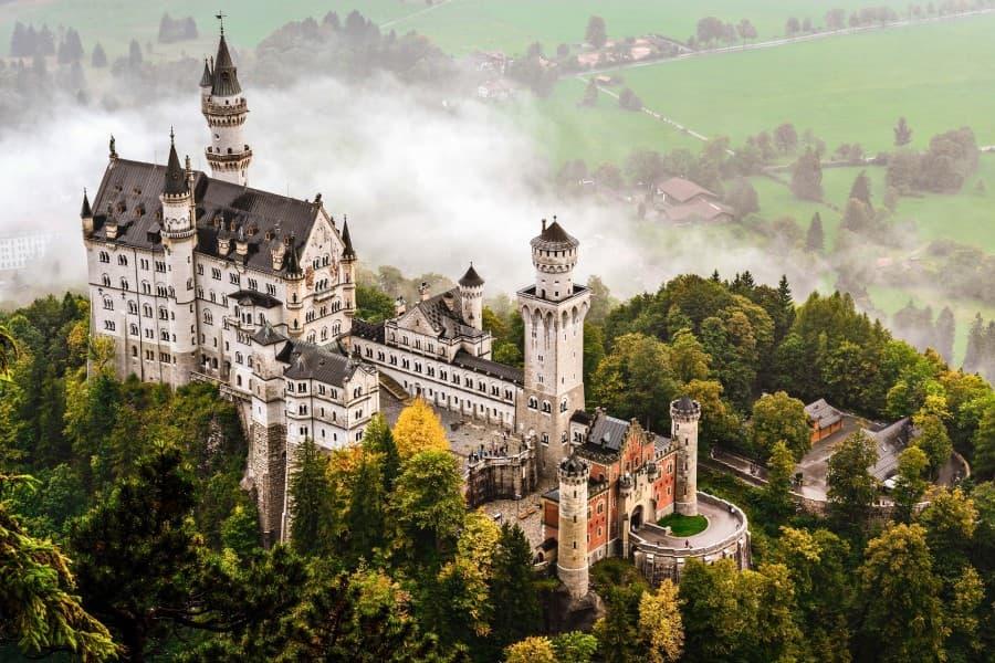 Vista aérea do Castelo de Neuschwanstein. Ele está situado em uma montanha, cercada por uma floresta e névoa, e se destaca pelos altos torres e pela impressionante arquitetura.