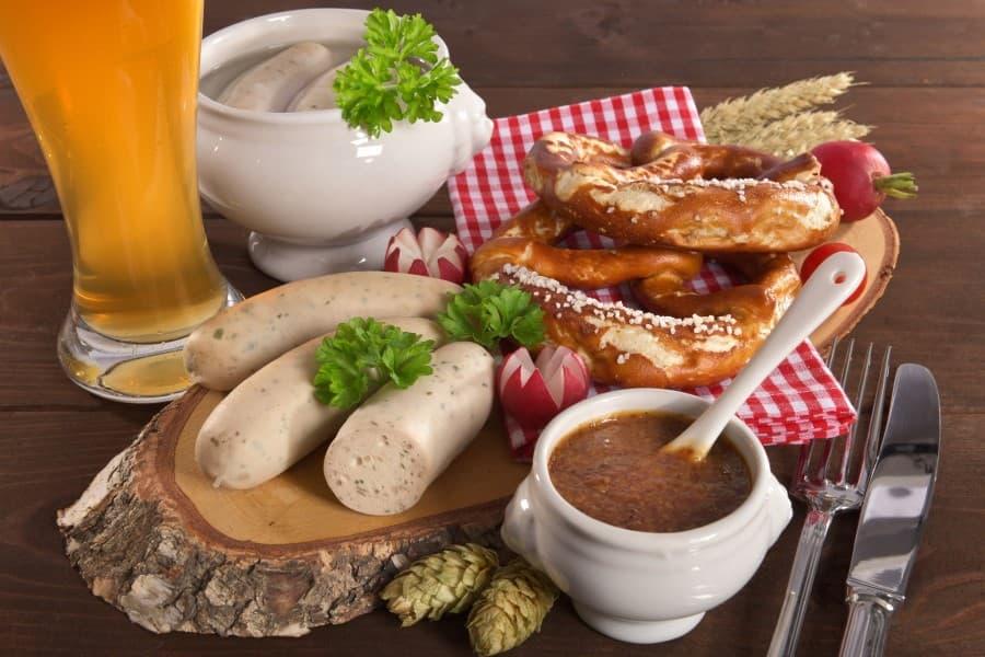 Delícias bávaras: um copo de cerveja de trigo, ao lado uma tigela branca com salsichas brancas. À frente, uma tábua de madeira usada como bandeja, onde estão três salsichas brancas e dois pretzels. Na frente da bandeja há também um pote de mostarda doce.