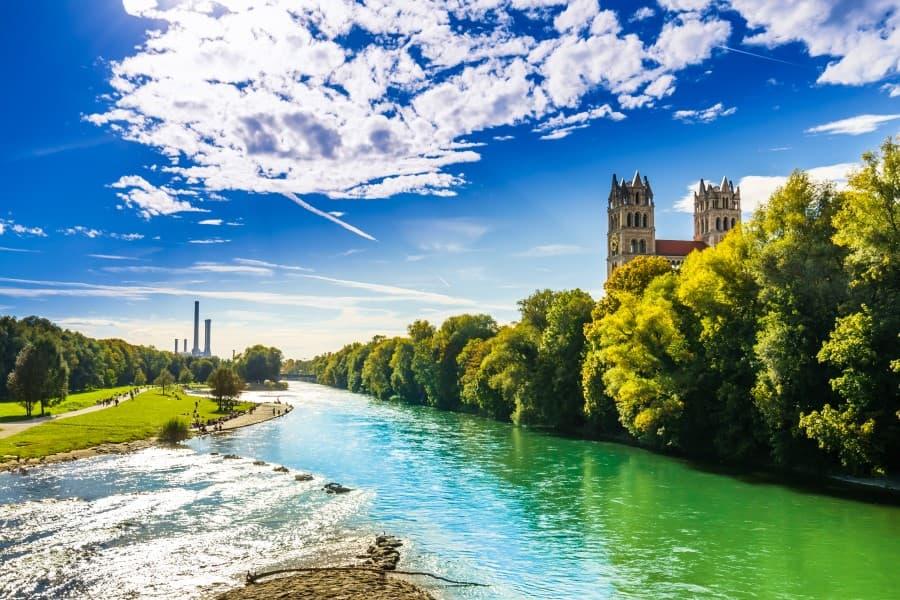 O rio Isar flui pelo centro da imagem, brilhando ao sol de verão sob o céu azul em tons de verde e azul. À direita, há muitas árvores verdes e arbustos, atrás se ergue um castelo com duas torres. À esquerda do rio, há um gramado entrecortado por um caminho para pedestres e ciclistas.