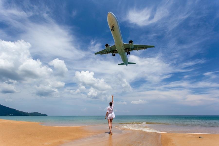 An airplane flies low over a sunny beach with a blue sky and clouds. A person in the foreground stands by the water looking up at the airplane that is flying directly above them.